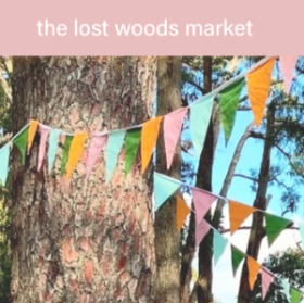 Image of a tree trunk in foreground with trees in the distance at The Lost Woods Market in Emerald at Puffing Billy Park. There is colourful bunting hanging between the trees and a pink banner is at the top of the image with the lost woods market in white text across it.